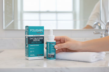 Tangan memegang serum DHT FOLIGAIN di sebelah kotaknya dan tuala berlipat di atas kaunter bilik mandi.