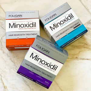 Tiga formula minoksidil FOLIGAIN dalam kotak jingga, biru dan ungu.