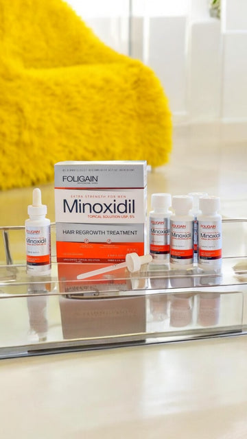 Kotak dan botol minoksidil FOLIGAIN di rak bilik mandi, di sebelah tuala kuning.