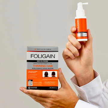 Tangan memegang kotak dan botol semburan rawatan sapuan TRIOXIDIL 10% FOLIGAIN untuk lelaki.
