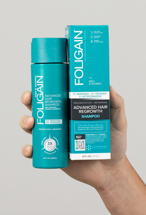 Tangan memegang syampu pertumbuhan semula rambut lanjutan FOLIGAIN dalam botol hijau kebiruan di sebelah kotaknya.